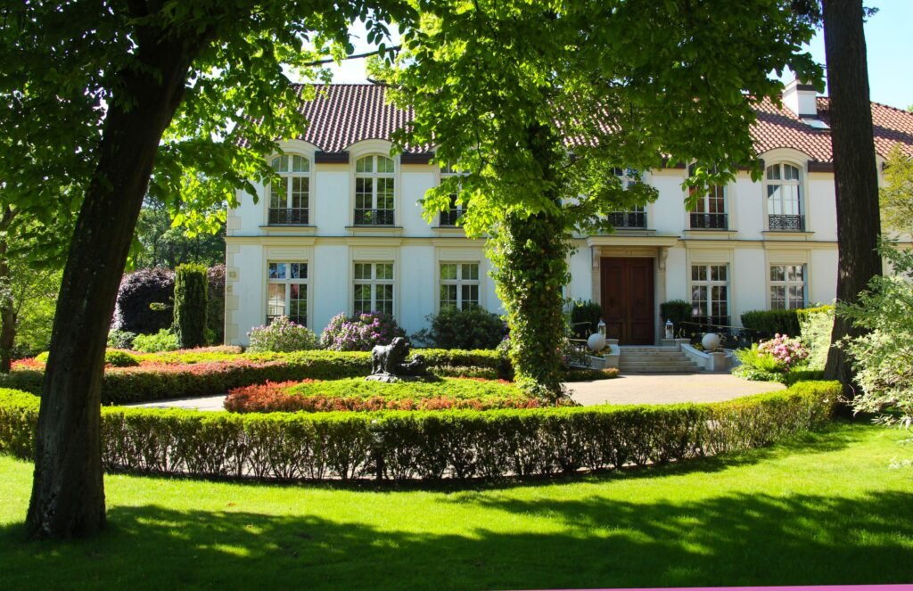 Stately white mansion with manicured gardens and lush trees under a bright sky.